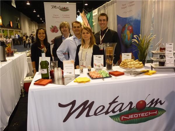 Five people stand behind a table with a white cloth bearing the Metarom Neotech logo. The table holds a wine bottle, glasses, small plates of food and signs. A Metarom banner stands behind them.