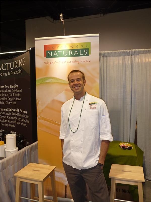 A smiling man in a white chef's coat and green bead necklace stands in front of a Northwest Naturals banner. Two wooden stools and a green draped table are nearby.