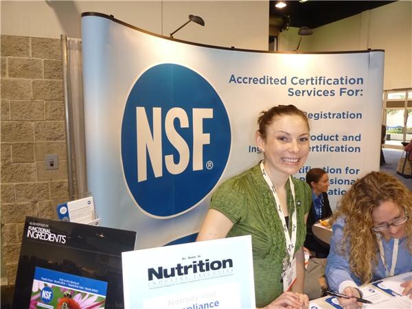 A smiling woman in a green top stands at the NSF booth with a large blue NSF logo behind her. A Nutrition Industry Executive sign and a Functional Ingredients display are in front. A seated woman is at right.