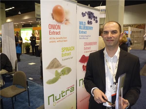 A man in a dark blazer with a lanyard holds brochures in front of Nutra Canada banners for onion, spinach, wild blueberry and cranberry extracts.