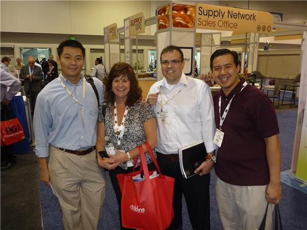 Four smiling people with lanyards pose together in front of a Supply Network Sales Office sign. A woman holds a red Chiptons tote bag, and a P&G bag is visible at left.
