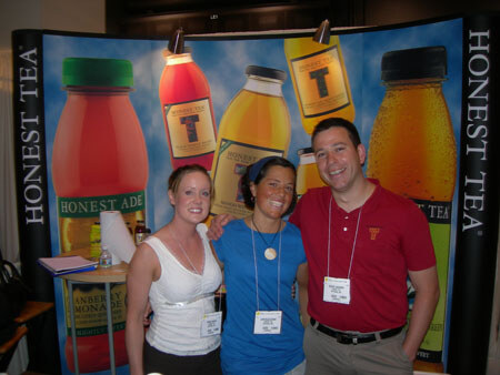 Two women and a man stand together in front of an Honest Tea display with large Honest Ade and tea bottle graphics.