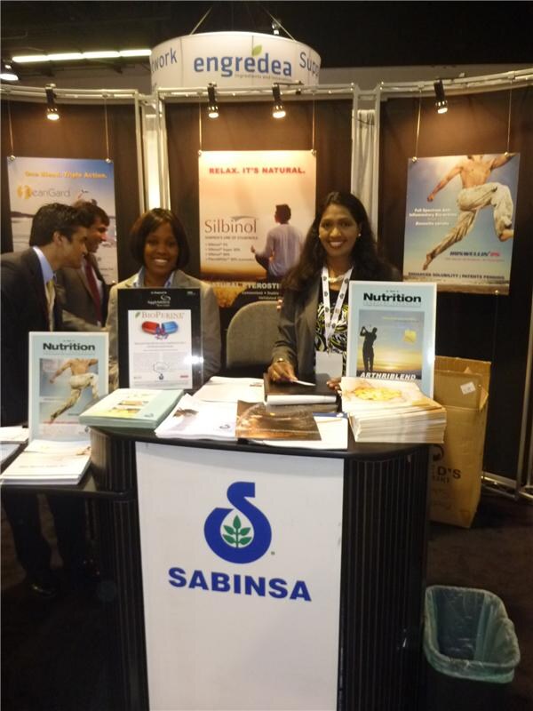 Two women stand smiling behind a Sabinsa counter with Nutrition magazine and BioPerine materials. Two men in suits stand at left. Silbinol and Boswellin posters and an Engredea sign are behind them.