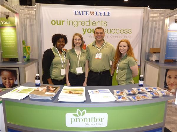 Four people in green polo shirts stand behind a curved Tate & Lyle booth counter with a Promitor Dietary Fiber logo, brochures and small snack packets on top.