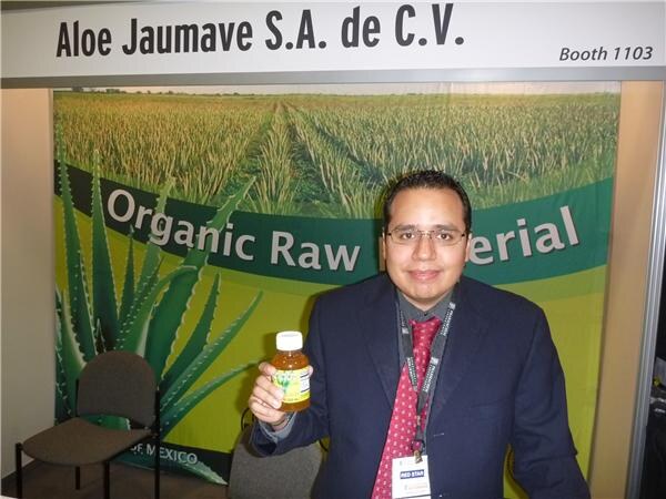 A man in a dark suit and red tie holds a small bottle of aloe juice in front of an Aloe Jaumave S.A. de C.V. Booth 1103 sign and an organic raw material banner.