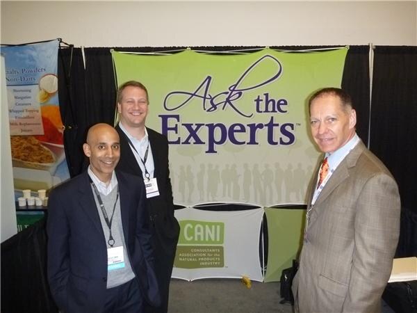 Three men in suits stand in front of an Ask the Experts banner with a CANI logo, the Consultants Association for the Natural Products Industry.