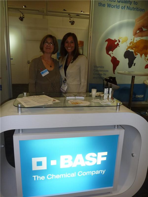 Two women stand behind a white curved BASF counter with a lit BASF The Chemical Company sign; a world map banner mentions vitamins, carotenoids and omega-3s.