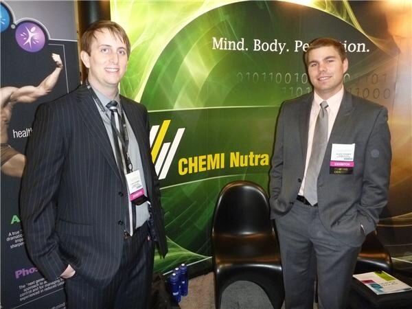 Two men in suits stand in front of a green Chemi Nutra backdrop with the slogan Mind. Body. Perf... partly visible; both wear exhibitor badges.
