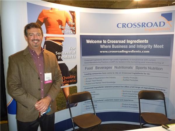 A man in a brown blazer stands beside a Crossroad Ingredients display listing food, beverage, nutritional and sports nutrition ingredients, with two folding chairs nearby.