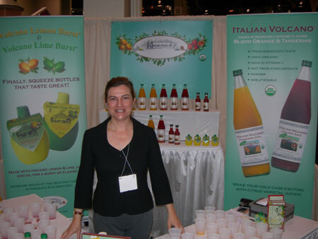 A woman with a name badge stands at a booth with banners for Italian Volcano juices and Volcano Lemon Burst and Lime Burst, with juice bottles on a table behind her.