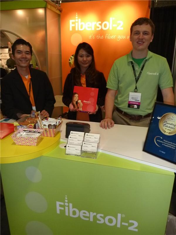 Two men and a woman stand behind a green and yellow Fibersol-2 counter; the woman holds an orange brochure, and snacks, business cards and an award plaque are displayed.