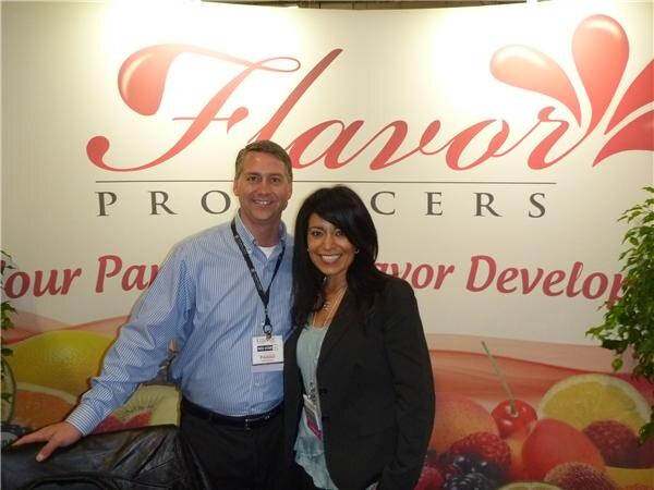 A man in a blue striped shirt and a woman in a black blazer pose in front of a Flavor Producers backdrop with a fruit graphic.