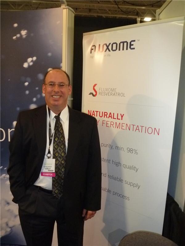 A smiling man in a dark suit and exhibitor badge stands with a hand in his pocket beside a Fluxome Resveratrol banner reading Naturally by fermentation.