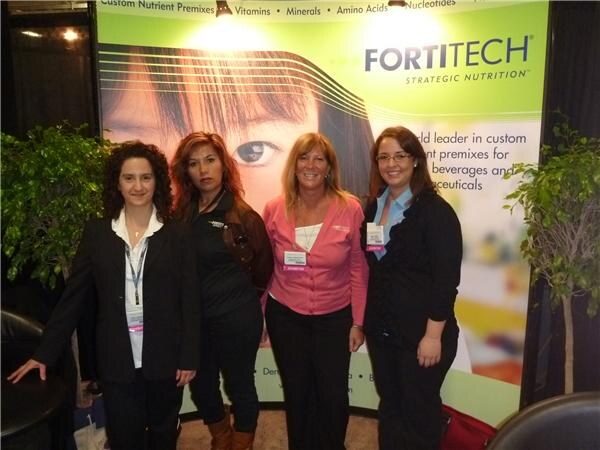 Four women stand side by side in front of a green Fortitech Strategic Nutrition backdrop showing a woman's face, with potted plants on both sides.