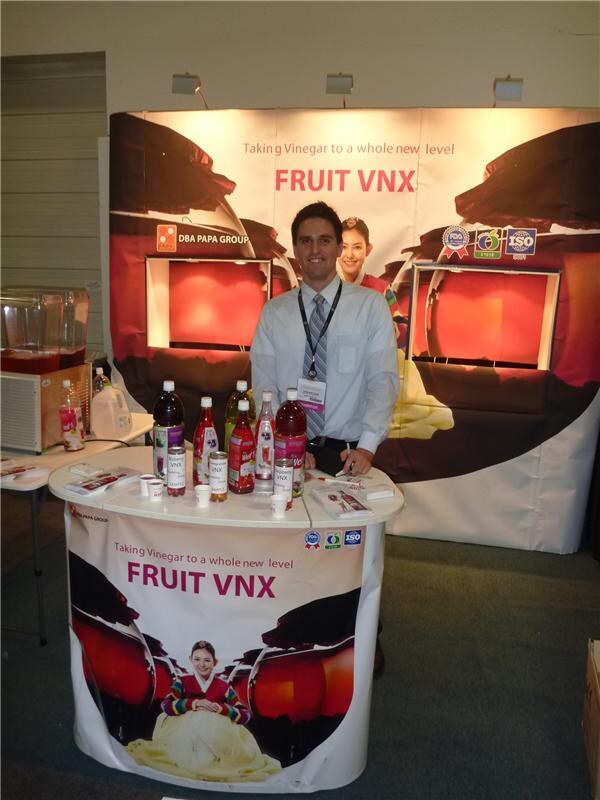 A man in a white shirt and tie stands behind a round counter holding bottles of Fruit VNX drinks, with Fruit VNX and DBA Papa Group signage behind him.