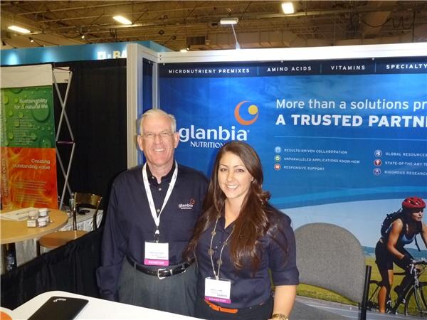 A man and a woman in dark navy shirts and exhibitor badges stand together in front of a blue Glanbia Nutritionals banner reading More than a solutions provider.