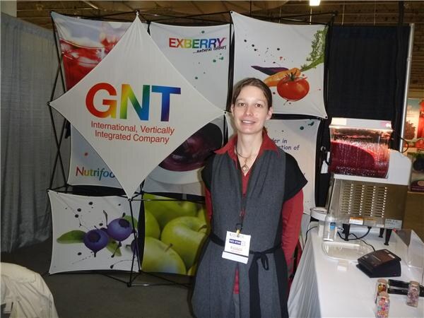 A woman in a gray vest over a red top stands in front of GNT and Exberry banners with fruit and vegetable graphics, next to a table with a display unit.
