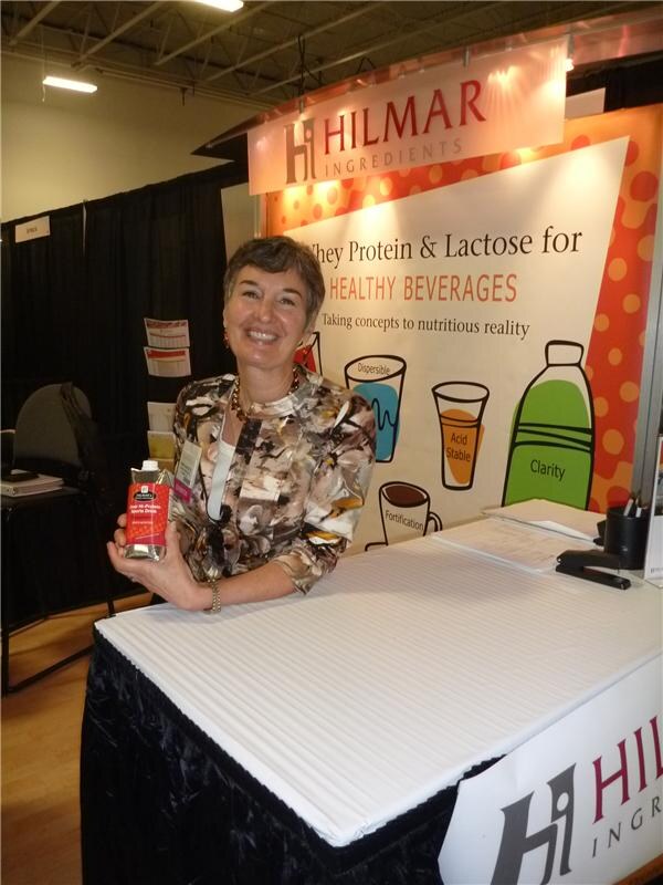 A smiling woman leans on a draped table holding a small carton drink, in front of a Hilmar Ingredients sign about whey protein and lactose for healthy beverages.