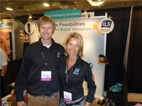 A man and a woman in black ILS shirts and exhibitor badges smile together in front of an ILS Innovative Labeling Solutions sign.