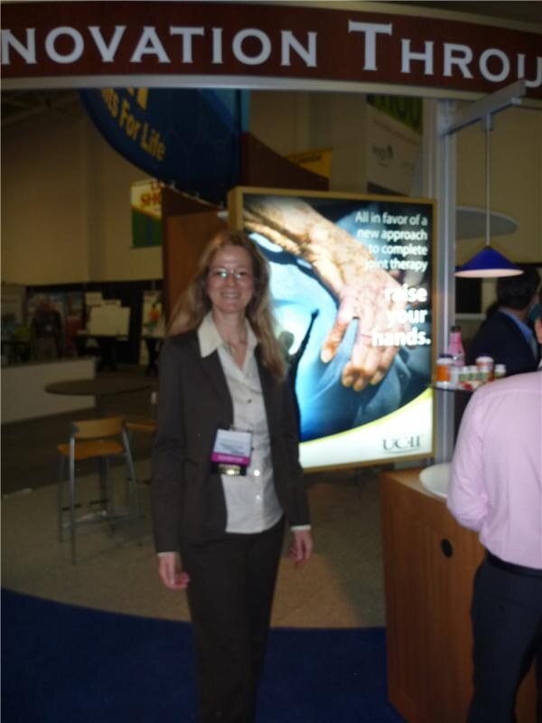 A woman with glasses and an exhibitor badge stands in front of a lit UC-II display reading Raise your hands, with another person at a counter at right.