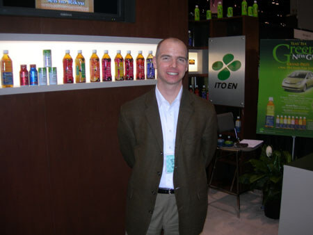 A smiling man in a brown blazer and white shirt stands in front of an ITO EN display with lit shelves of bottled tea and a green ITO EN sign.