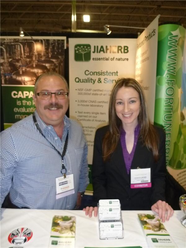 A man with glasses and a mustache and a woman in a dark blazer stand behind a table with business cards and brochures, in front of Jiaherb banners.