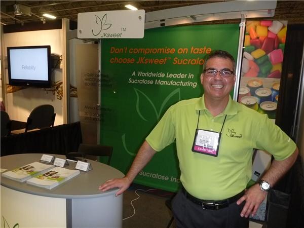 A smiling man in a green polo shirt and glasses leans on a round table in front of a green JKsweet sucralose banner with a candy image.