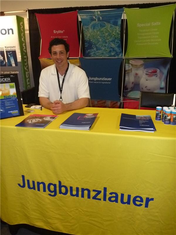 A smiling man in a white polo and lanyard sits behind a yellow Jungbunzlauer table holding brochures, with Erylite and Special Salts banners behind him.