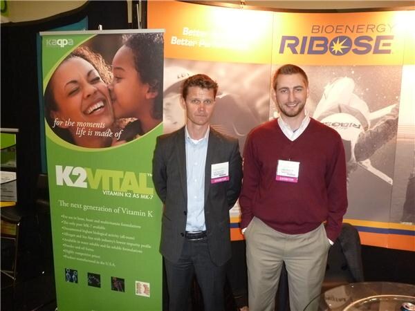 Two men with exhibitor badges stand between a green K2Vital banner on the left and a Bioenergy Ribose display on the right.