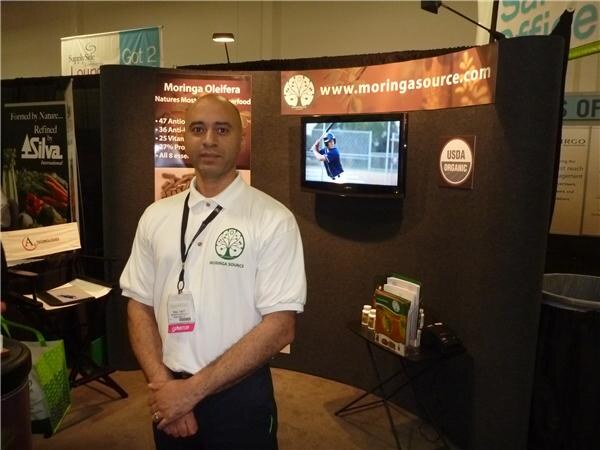 A bald man in a white Moringa Source polo and lanyard stands before a booth with a wall-mounted screen, a USDA Organic seal and the moringasource.com web address.
