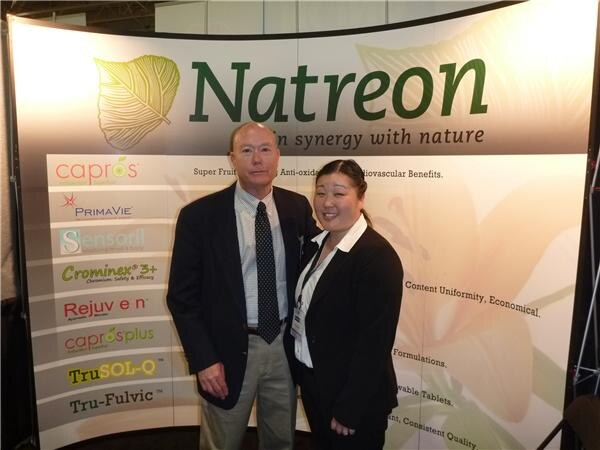 A man and a woman in dark clothes stand in front of a Natreon backdrop listing Caprōs, PrimaVie, Sensoril, Crominex 3+, Rejuven, Caprōsplus, TruSOL-Q and Tru-Fulvic.