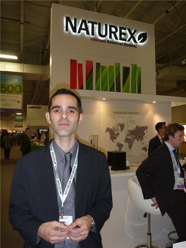 A man in a pinstripe jacket and Naturex lanyard stands before a lit Naturex sign and an industrial network map; a Lonza sign and other attendees appear behind.