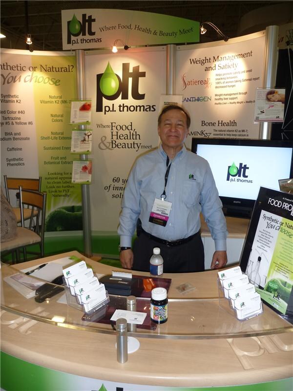 A smiling man in a light blue shirt stands behind a curved counter with business card holders at the p.l. thomas booth, with Satiereal and Anthigen signs behind.