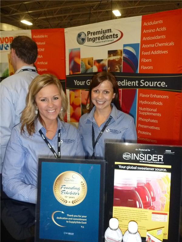 Two smiling women in striped shirts and lanyards stand at the Premium Ingredients International booth, beside a Founding Exhibitor plaque and a Natural Products Insider ad.
