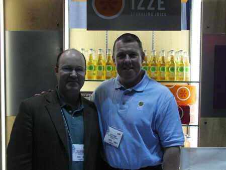 Two men stand side by side with arms around each other in front of shelves of Izze sparkling juice bottles under an Izze sign.