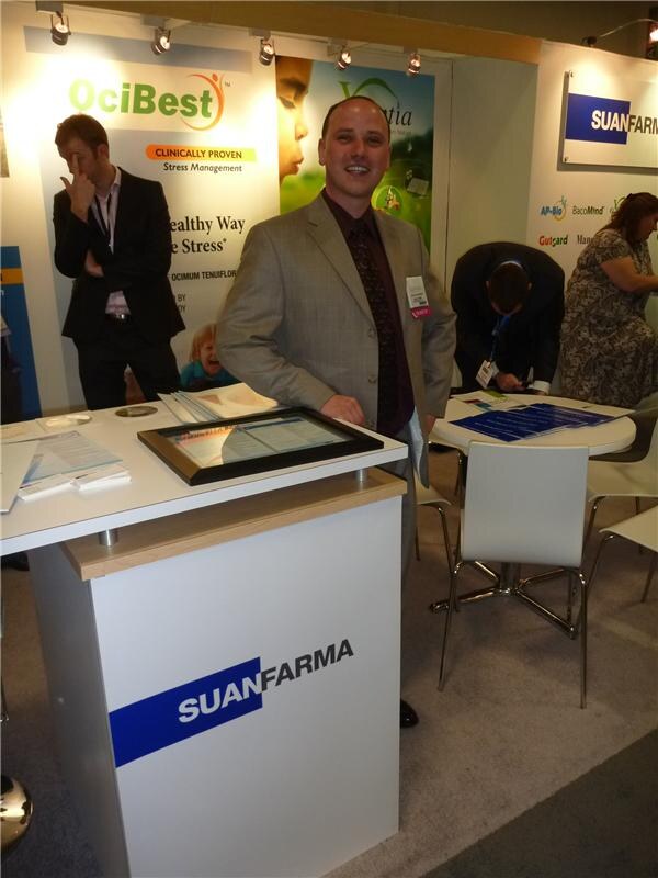 A smiling man in a gray jacket stands beside a Suanfarma counter; OciBest, AP-Bio, BacoMind and Gutgard signs are behind him, with other attendees nearby.