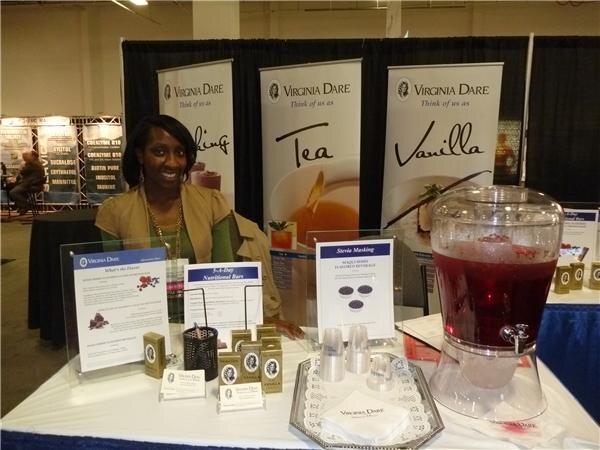 A smiling woman in a tan jacket sits at a Virginia Dare booth with banners for Tea and Vanilla, product cards, small boxes and a dispenser of red iced tea on a white tablecloth.