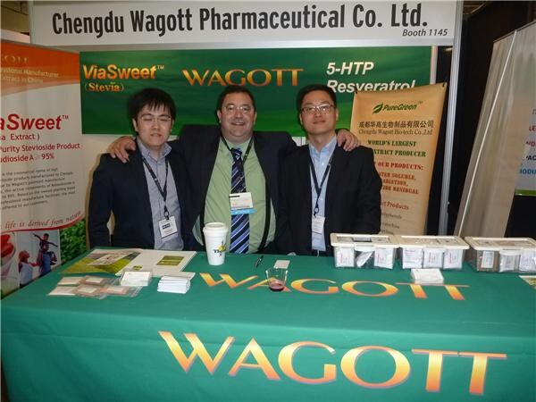Three men in suits stand with arms around each other behind a green Wagott table under a Chengdu Wagott Pharmaceutical Co. Ltd. sign, with ViaSweet and PureGreen signage behind them.