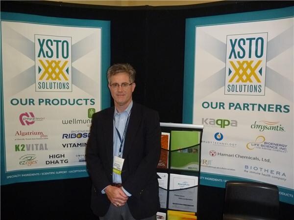 A man with glasses and a dark blazer stands between two XSTO Solutions banners listing products such as Pepzinor, Algatrium Plus and K2Vital and partners including Kaqpa, Chrysantis and Biothera.