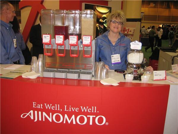 A smiling woman with glasses in a blue shirt stands behind a red Ajinomoto counter reading Eat Well, Live Well, with a four-tank beverage dispenser, cups and napkins; a man stands at left.