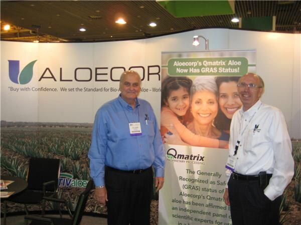 Two men in button-up shirts and badges stand in the Aloecorp booth beside a Qmatrix banner announcing GRAS status for Aloecorp's Qmatrix aloe, with an aloe field backdrop behind them.
