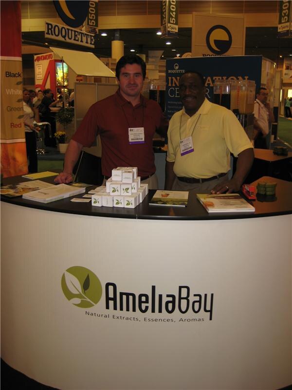 Two men in a red polo and a yellow polo stand behind a curved white Amelia Bay counter holding stacked small boxes and brochures, with other booths visible behind them.
