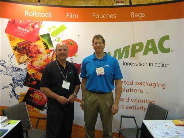 Two men stand in front of an orange and white Ampac backdrop that lists rollstock, film, pouches and bags and shows food packaging images; one wears a black shirt and the other a blue polo.