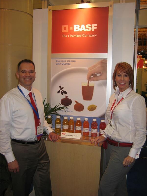 A man in a white shirt and red tie and a woman with red hair in a white shirt stand beside a BASF display with a row of bottled orange and red beverages on a wooden counter.