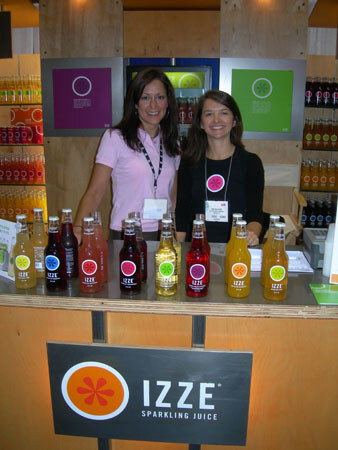 Two smiling women stand behind a counter lined with colorful Izze sparkling juice bottles, with an Izze sign on the front of the counter.