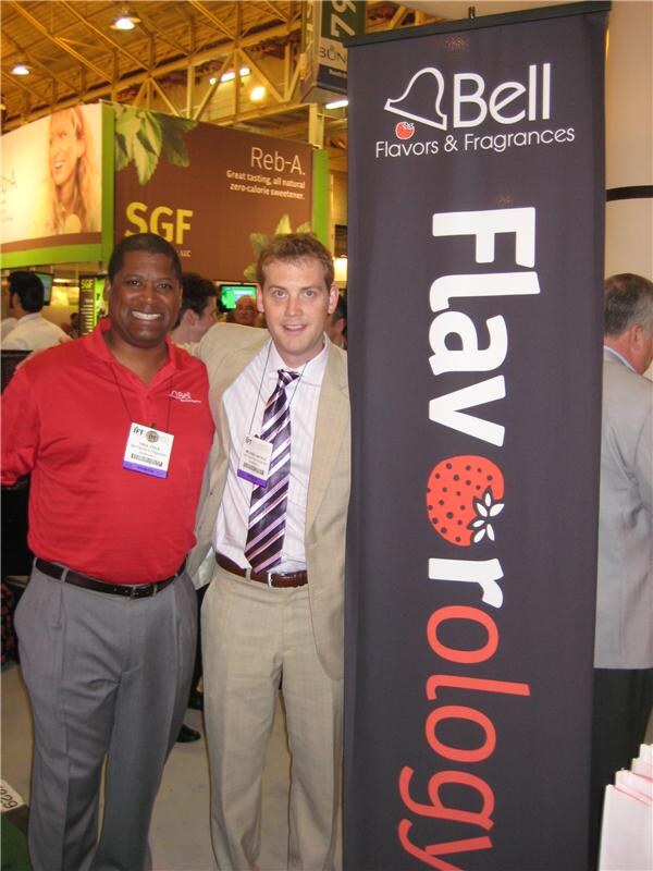 Two men stand beside a tall dark Bell Flavors & Fragrances banner reading Flavorology; one wears a red polo and the other a tan suit with a striped tie. An SGF sign is behind them.