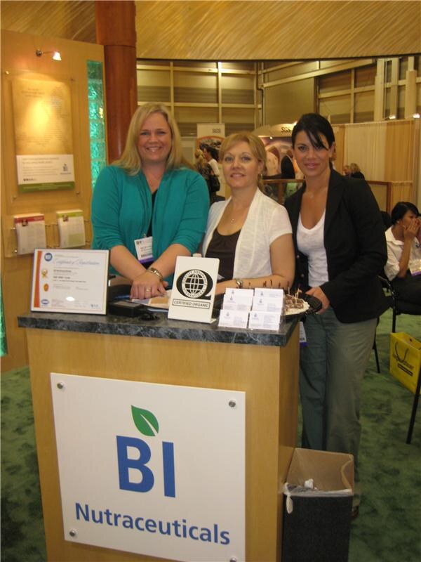 Three women stand behind a wooden counter at the BI Nutraceuticals booth, with business cards, a certified organic sign and a framed certificate on the counter.