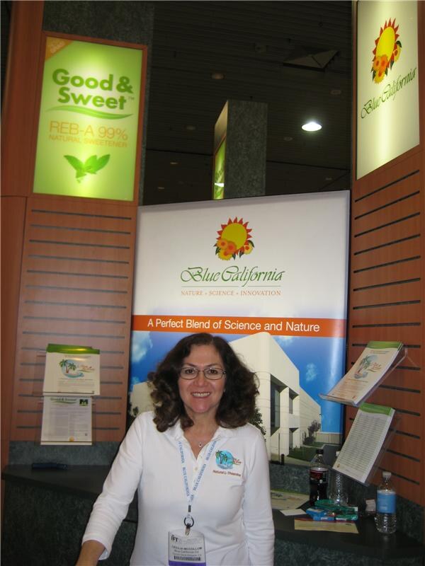 A smiling woman with curly hair and glasses stands in the Blue California booth, in front of a Blue California banner and a Good & Sweet REB-A 99% natural sweetener sign.