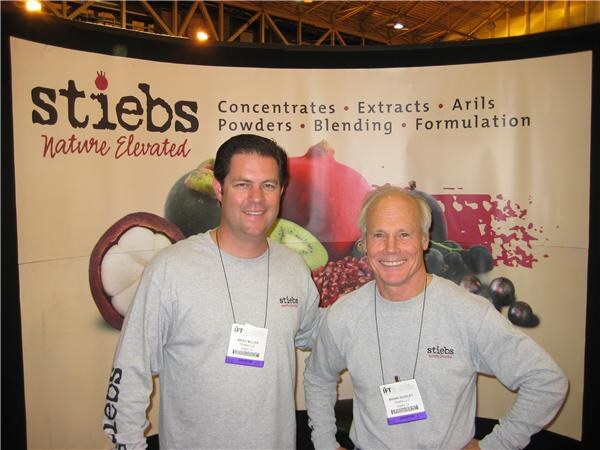Brad Miller (left) and Brian Dudley (right) wear gray Stiebs long-sleeve shirts and badges, standing in front of a Stiebs banner showing pomegranate, kiwi and mangosteen.