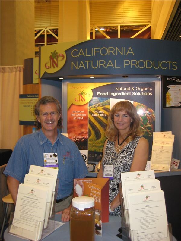 A man in a denim shirt and a woman in a patterned dress stand at the California Natural Products booth, with brochures and a jar of syrup on the counter in front of them.